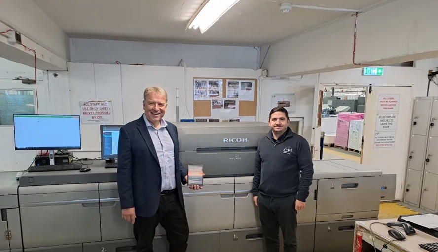 Two men smiling in a printing facility, standing in front of a large Ricoh printer. One holds a printed item. The space is industrial and organized.