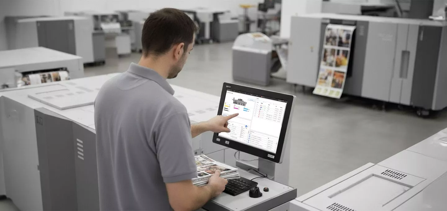  A person is standing in a print production facility, using a touchscreen computer to control a large Ricoh printer. The person is pointing at the screen, which displays a complex user interface with various settings, charts, and diagrams. The person is holding a stack of printed materials, including a brochure. Multiple industrial printers are visible in the blurred background.