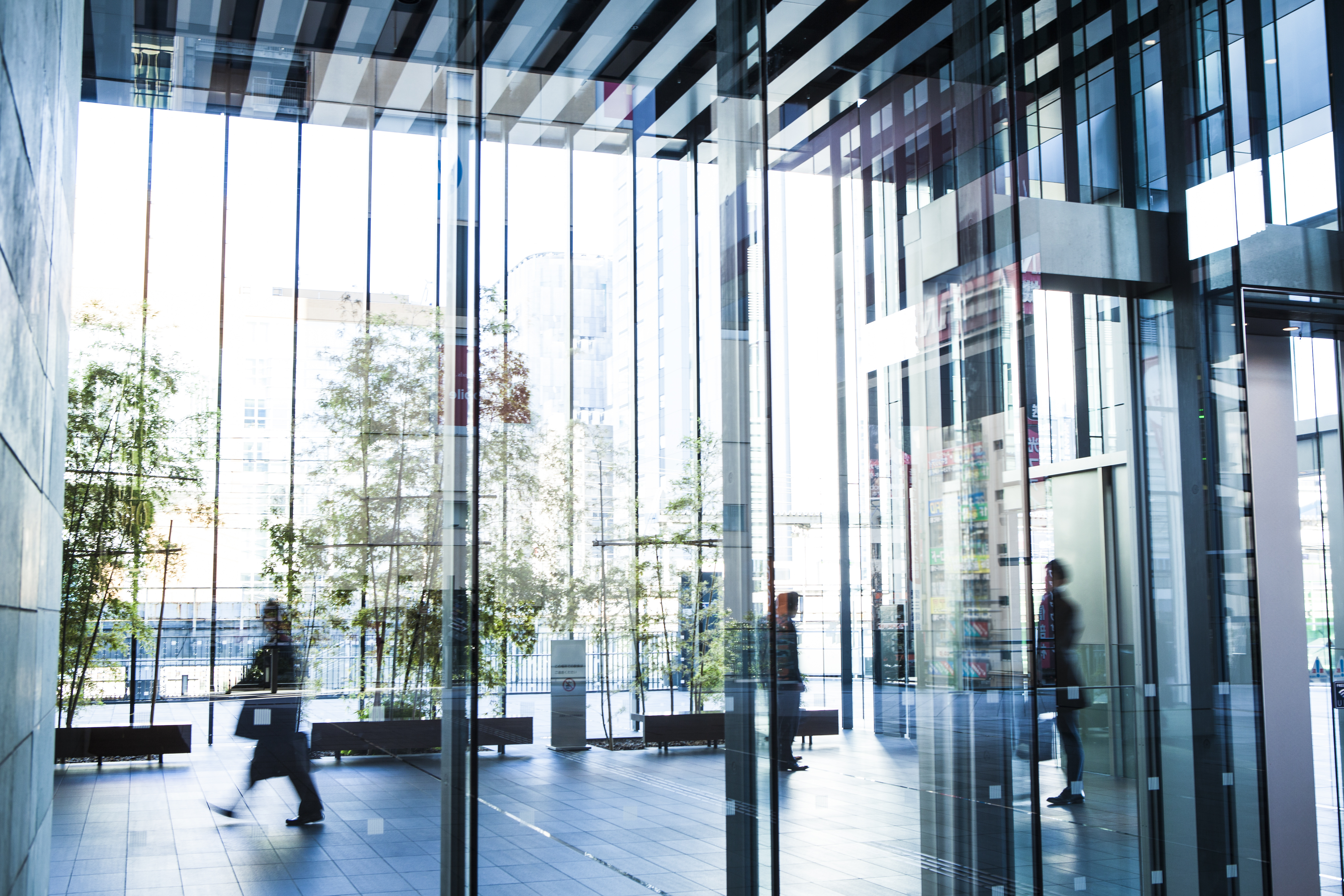 Photograph of a modern office building lobby with large glass walls and reflections creating a layered effect. Several blurred figures of people walking and standing are visible, emphasizing movement and activity within a sleek, urban environment.
