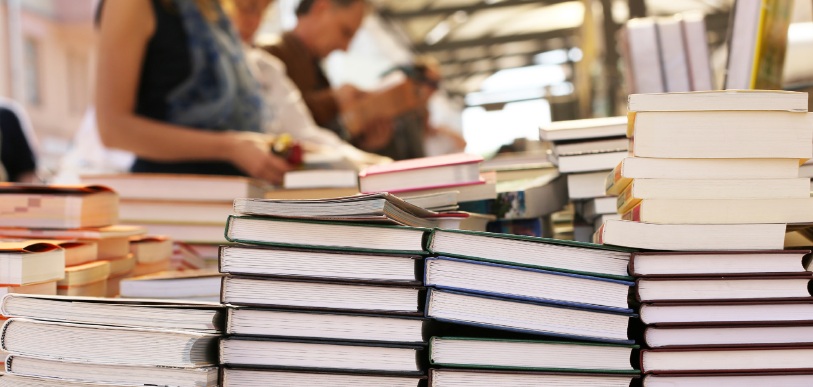 Stacks of books displayed on an outdoor market stall, with blurred people browsing in the background. The scene conveys a lively, bustling atmosphere.